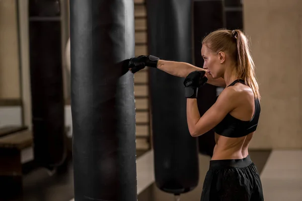 Atleta rubia hembra con auriculares saco de boxeo con guantes de boxeo en el gimnasio —  Fotos de Stock