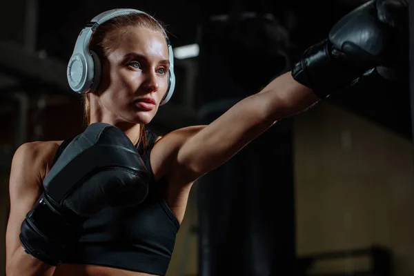 Atleta rubia hembra con auriculares saco de boxeo con guantes de boxeo en el gimnasio — Foto de Stock