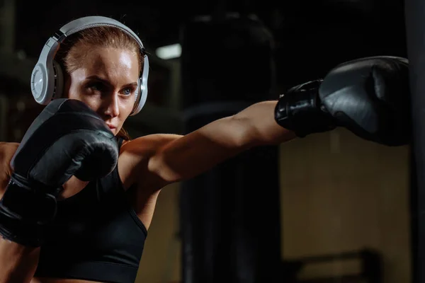 Atleta rubia hembra con auriculares saco de boxeo con guantes de boxeo en el gimnasio — Foto de Stock