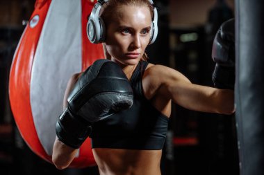 Kulaklıklı sarışın bir atlet. Spor salonunda boks eldivenli bir kum torbası.