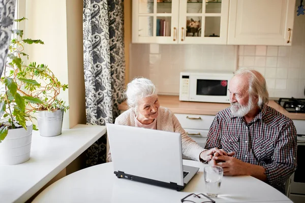 Encantador casal sênior usando laptop sentado à mesa na sala de cozinha. — Fotografia de Stock