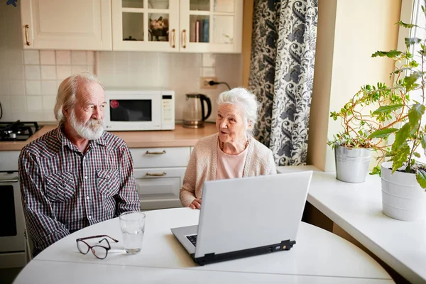 Affascinante coppia anziana utilizzando il computer portatile seduto al tavolo in cucina. — Foto Stock