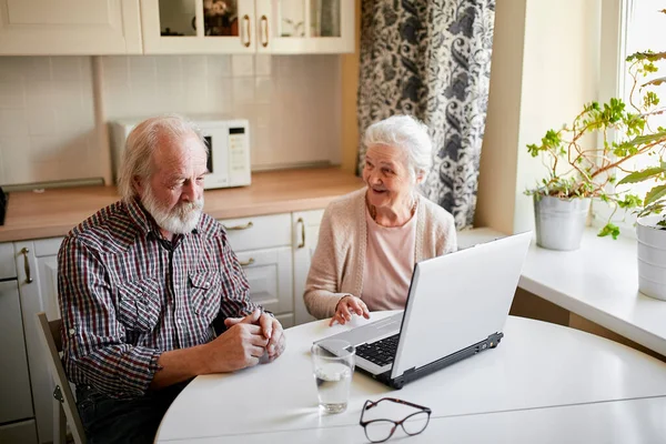 Encantadora pareja de personas mayores utilizando el ordenador portátil sentado en la mesa en la sala de cocina. — Foto de Stock