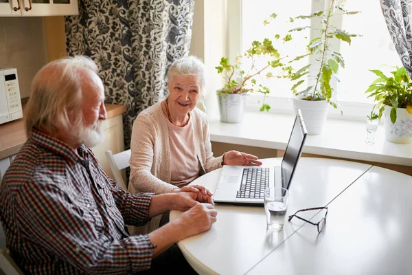 Affascinante coppia anziana utilizzando il computer portatile seduto al tavolo in cucina. — Foto Stock