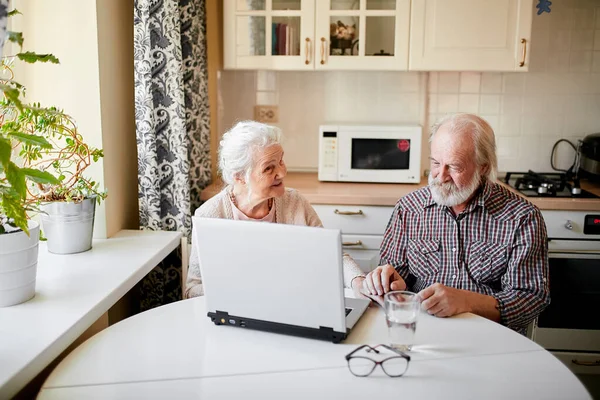 Encantador casal sênior usando laptop sentado à mesa na sala de cozinha. — Fotografia de Stock