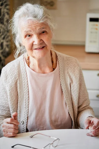 Portrét krásné starší ženy usmívající se před kamerou, sedící doma u stolu. — Stock fotografie