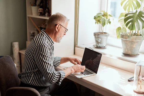 Mature bearded man working from home with laptop. sitting at desk near window — Stock Photo, Image