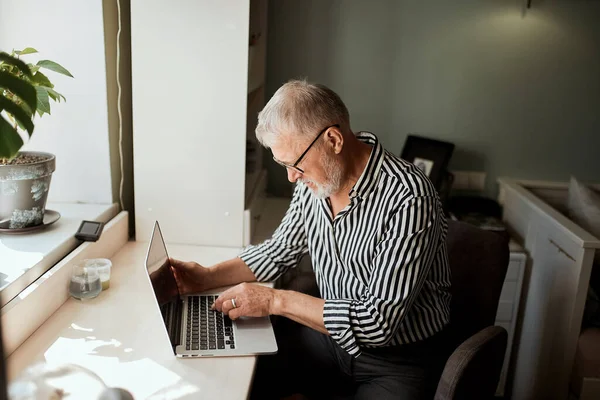 Dospělý vousatý muž pracující z domova s laptopem. posezení u stolu u okna — Stock fotografie
