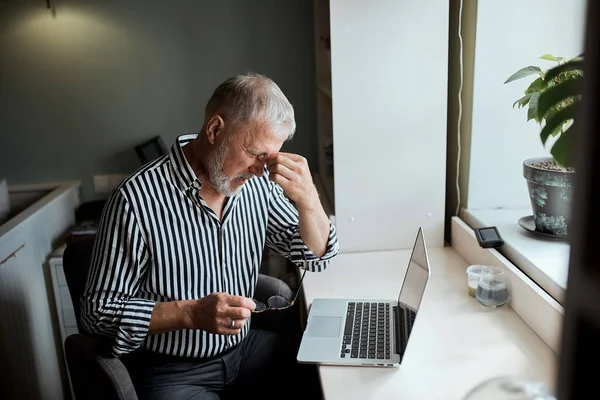 Hombre maduro mirando agotado mientras está sentado en su computadora portátil y sosteniendo sus gafas en la mano — Foto de Stock