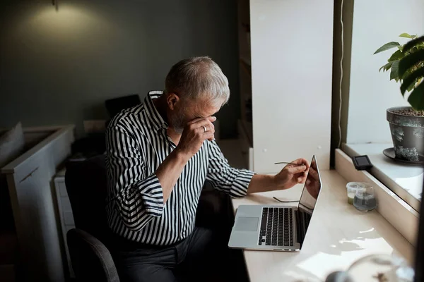 Hombre maduro mirando agotado mientras está sentado en su computadora portátil y sosteniendo sus gafas en la mano — Foto de Stock