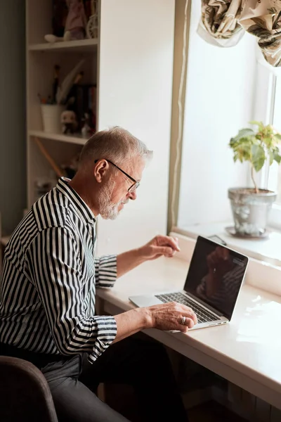 Ώριμος γενειοφόρος άνδρας που εργάζεται από το σπίτι με φορητό υπολογιστή. κάθεται στο γραφείο κοντά στο παράθυρο — Φωτογραφία Αρχείου