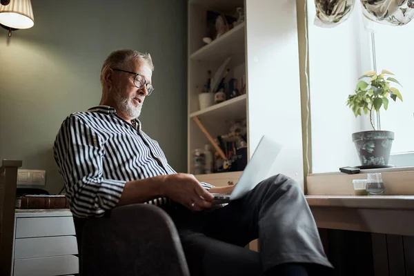 一个成熟的留着胡子的男人带着笔记本电脑在家里工作。坐在靠窗的桌子旁边 — 图库照片