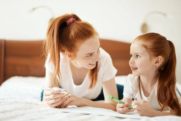 Mère aux cheveux roux se couche sur le lit avec sa fille d'âge préscolaire et dessine. — Photo