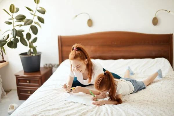 Kızıl saçlı anne, anaokulu kızıyla yatağa uzanıyor ve resim çiziyor.. — Stok fotoğraf