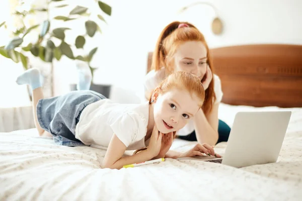 Pelirroja mamá y su hija tumbado en la cama en casa mientras mira el ordenador portátil —  Fotos de Stock