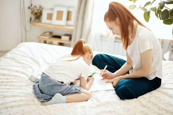 Kızıl saçlı anne, anaokulu kızıyla yatağa uzanıyor ve resim çiziyor.. — Stok fotoğraf