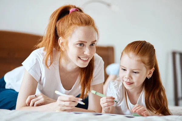 Mère aux cheveux roux se couche sur le lit avec sa fille d'âge préscolaire et dessine. — Photo