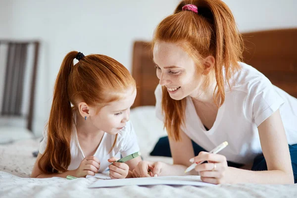 Mère aux cheveux roux se couche sur le lit avec sa fille d'âge préscolaire et dessine. — Photo