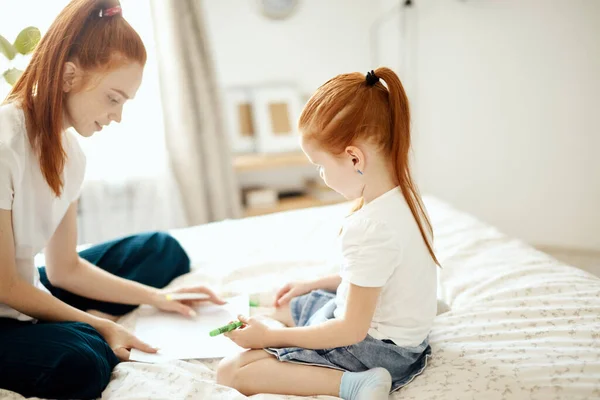 Mère aux cheveux roux se couche sur le lit avec sa fille d'âge préscolaire et dessine. — Photo