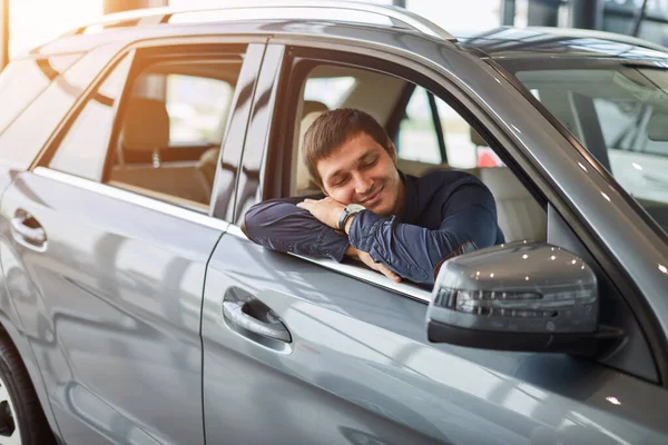 Посещение автосалона. Мечтающий мужчина обнимает новую машину и улыбается — стоковое фото