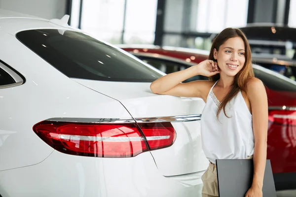 Female seller holding black folder looking at camera at car dealership.