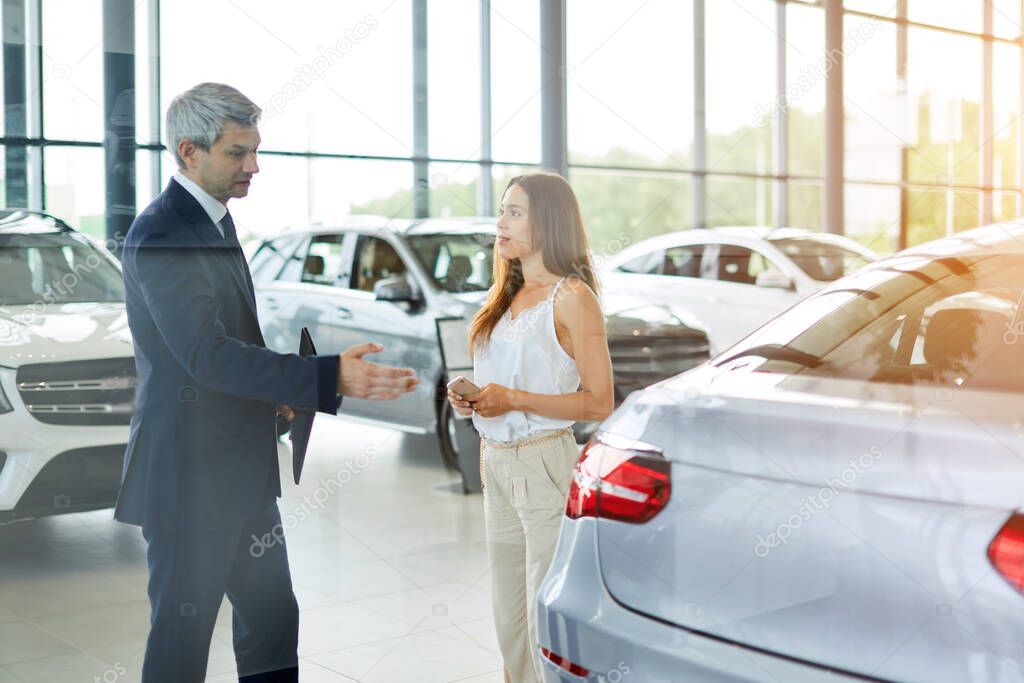 Young female customer asking mature sales manager to help in car dealership.