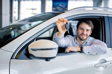 Araba galerisindeki yeni arabada oturan neşeli genç adam anahtarı tutuyor.