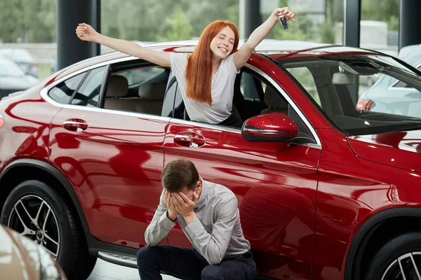 Щаслива жінка святкує покупку своєї нової дорогої машини, поки чоловік розчарований — стокове фото