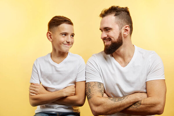 father and cute son looking at each other and smiling, crossed arms, yellow background
