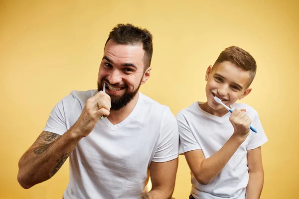 Pai e filho escovando os dentes e olhando para a câmera juntos isolados no amarelo — Fotografia de Stock