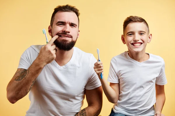 Padre e hijo cepillándose los dientes y mirando a la cámara juntos aislados en amarillo — Foto de Stock