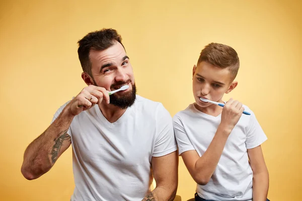Батько і син чистять зуби і дивляться на камеру разом ізольовані на жовтому — стокове фото