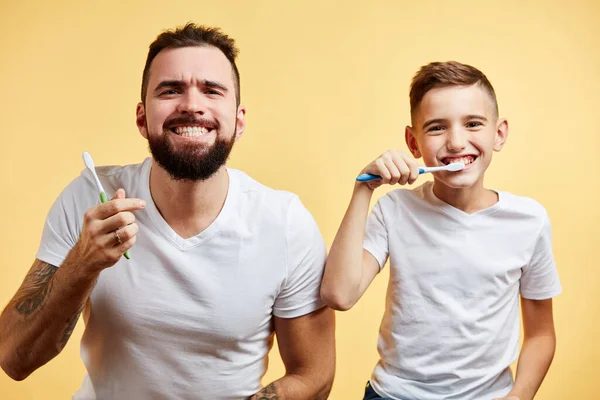 Padre e hijo cepillándose los dientes y mirando a la cámara juntos aislados en amarillo — Foto de Stock