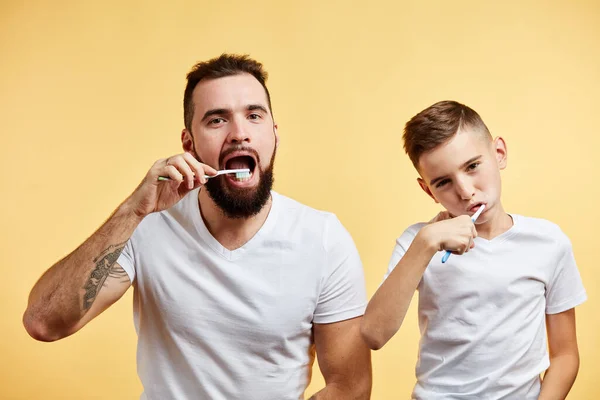 Padre e hijo cepillándose los dientes y mirando a la cámara juntos aislados en amarillo — Foto de Stock