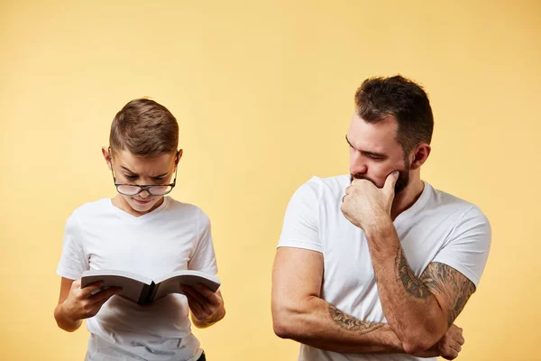Padre incredulo guardando carino piccolo figlio lettura libro su sfondo giallo — Foto Stock