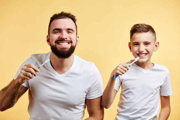 father and son brushing teeth and looking at camera together isolated on yellow