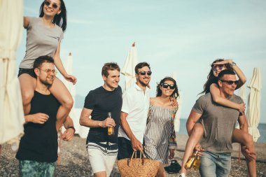 Bir grup arkadaş sahilde yürüyor, eğleniyor, kadınlar erkekleri sırtında taşıyor, komik bir tatil.