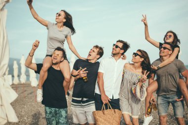 Bir grup arkadaş sahilde yürüyor, eğleniyor, kadınlar erkekleri sırtında taşıyor, komik bir tatil.