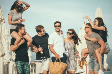 Bir grup arkadaş sahilde yürüyor, eğleniyor, kadınlar erkekleri sırtında taşıyor, komik bir tatil.
