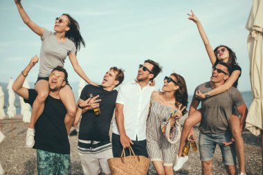 Bir grup arkadaş sahilde yürüyor, eğleniyor, kadınlar erkekleri sırtında taşıyor, komik bir tatil.