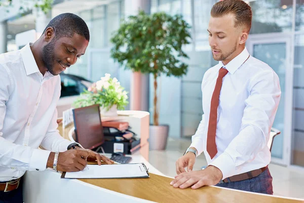 Acquirenti di auto. clienti africani che firmano documenti con il venditore nel negozio di concessionarie — Foto Stock