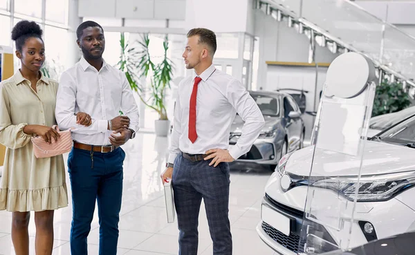 Schöner junger Autoverkäufer, der einem afrikanischen Paar von den Eigenschaften des Autos erzählt — Stockfoto