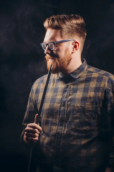 Stylový vousáč muž v brýlích a kostkované košili kouření hookah v nočním baru. — Stock fotografie
