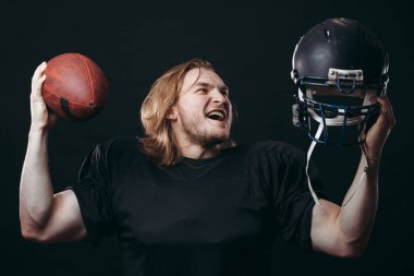 Siyah spor giyimli kendine güvenen Amerikan futbolcusu kask ve top tutuyor.