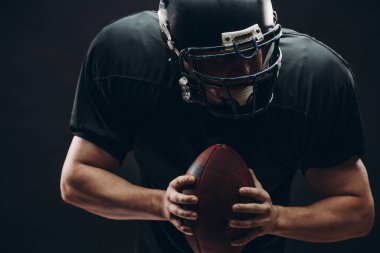 Kasklı ve zırhlı Amerikan futbolcu siyah duvara karşı top taşıyor.
