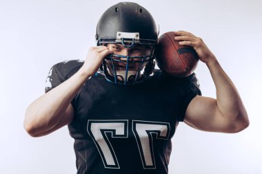 Koruyucu üniformalı ve kasklı Amerikan futbolcusu elinde top tutuyor.