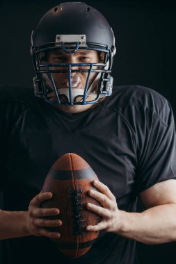 Siyah arka planda topu olan siyah spor giyimli Amerikalı bir futbolcu.