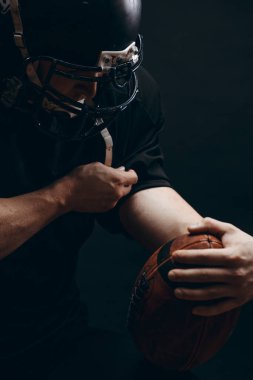 Siyah arka planda topu olan siyah spor giyimli Amerikalı bir futbolcu.