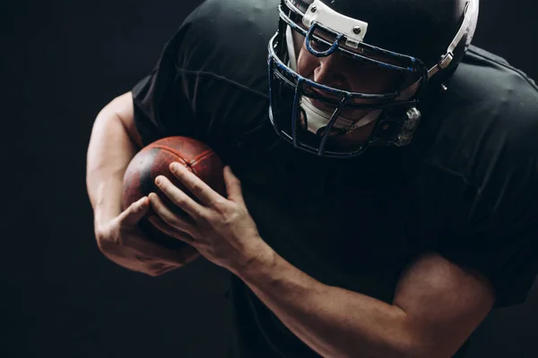 Jugador de fútbol americano con casco y armadura con una pelota contra la pared negra — Foto de Stock