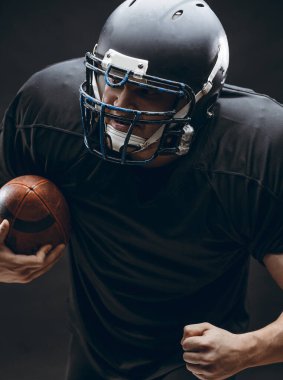 Kasklı ve zırhlı Amerikan futbolcu siyah duvara karşı top taşıyor.